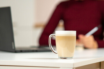 Glass mug of latte coffee in the foreground with a laptop and a woman writing in the background. Cozy remote work and studying atmosphere.