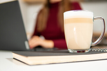 Close-up of a layered latte in a glass cup placed on a black notebook next to a laptop. Concept of business planning, studying, and coffee breaks.