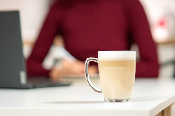 Freshly made layered cappuccino on a white desk. In the background, a woman in a red sweater is studying or working. Concept of focus and comfort.