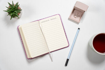 Wedding Plans notebook on a white desk with a diamond engagement ring, tea, and plant. Ideal for bridal blogs, event planners, and marriage content.