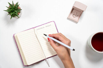 POV of a bride-to-be writing in a Wedding Plans journal. Features a diamond ring and tea, perfect for wedding organization concepts.