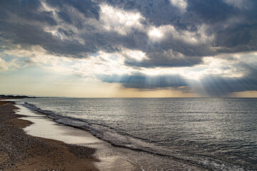 A deserted Mediterranean beach under a cloudy sky, where sunbeams break through the clouds to illuminate the calm sea. A peaceful interseason moment on the shore in Sorgun, Antalya, Turkey.

