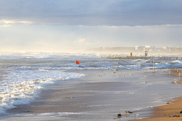 Stormy sea with massive waves and white foam under bright sunlight. Tilted volleyball poles stand in surf as red storm flag waves in the turbulent water. Side, Antalya, Turkey, Mediterranean.


