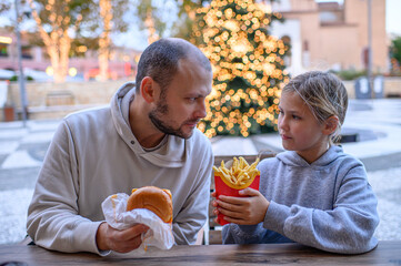 Father and daughter eating fast food in a shopping mall food court, enjoying a casual meal together and sharing a warm family moment indoors.