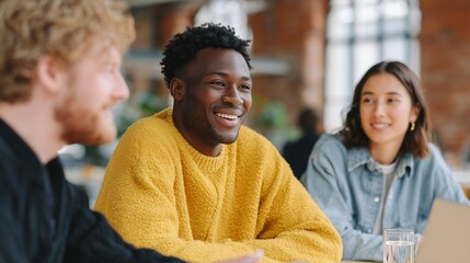 Smiling african american man in bright yellow sweater engages in creative business meeting with colleagues in sunlit industrial workspace, fostering collaboration and innovation