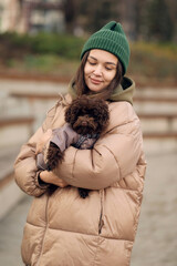 Young woman holding small poodle dog outdoors in autumn park, showing pet care, companionship and everyday lifestyle.