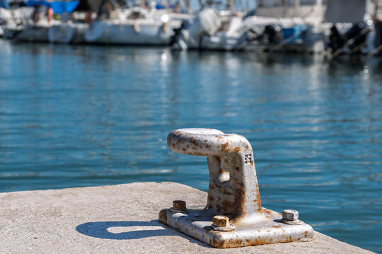 Close-up of an old weathered iron mooring bollard on a dock. Sunny day in a mediterranean marina with calm blue water and blurred white yachts in the background. Nautical detail and marine corrosion