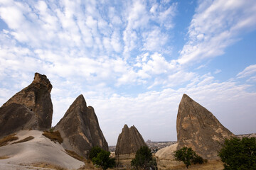 A scenic landscape featuring the unique rock formations and soft volcanic terrain of Cappadocia under a bright sky filled with scattered clouds.
