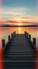 Obraz premium A wooden pier stretches out over a tranquil lake at sunset. The sky is a soft shade of orange and blue, with the reflection of the sunset light on the smooth surface of the water creating a peaceful a