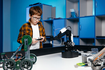 Young boy with glasses operates robotic arm in a modern classroom, surrounded by various robotics kits and tools, showcasing hands-on learning and innovation in technology