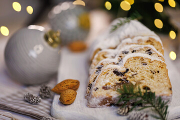 A close-up of a holiday stollen sliced and topped with powdered sugar, surrounded by almonds, pine sprigs, and festive ornaments on a rustic wooden display with holiday lights.