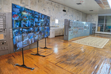 Hydroelectric Power Station control room. Screens on the left show hydroturbines, a large panel with gauges on the right. Parquet floor with a rug, marble walls.