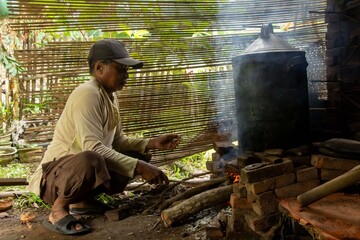 An old brown-skinned Asian man is boiling water with firewood in a simple kitchen with bamboo curtains in the background.