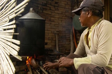 An Indonesian Asian man is cooking in the kitchen with firewood, the kettle looks black due to the smoke from the fire