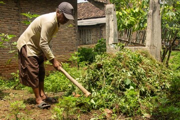 an old Asian man is cleaning the yard with his hoe with an outdoor background