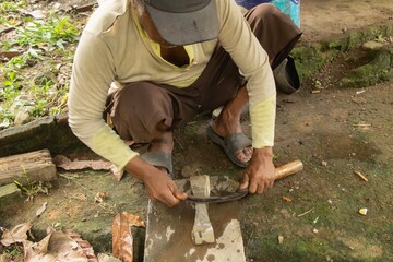 The appearance of an old Asian man wearing a dull hat with simple culture was rubbing a knife or machete on a pestle outside.