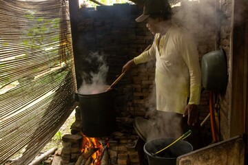 An old man wearing an Asian hat is boiling hot water in the kitchen using simple equipment typical of Indonesian culture.