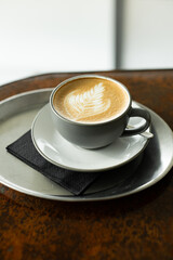 Coffee cup with intricate latte art on a saucer, black napkin, and metal tray on a rustic wooden cafe table