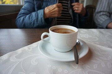 Weiße Tasse Kaffee mit silbernem Löffel auf weißer Stofftischdecke mit Muster vor altem Ehepaar in Café am Nachmittag im Herbst