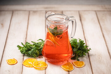 Red citrus lemonade in a glass pitcher with mint, orange slices and fresh herbs on rustic wooden background