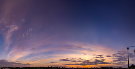 Sunset over calm river shore panoramic view. Dramatic sunset violet dark. Yellow Orange blue color shades cloudy sky. A strip of forest silhouette fills horizon line.