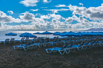 Serene view of a beach in the Canary Islands with dark sand and blue ocean. Rows of unoccupied blue...