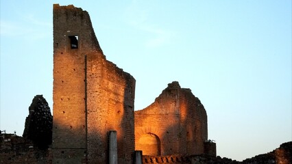 Ancient Roman ruins of a historical building or tomb stand partially lit by warm sunrise light against a clear pale blue sky, evoking antiquity and heritage along the Via Appia Antica
