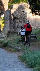 Adults and children cycling along the ancient cobblestone road, passing historical Roman ruins and lush green vegetation on a warm day, symbolizing family recreation and historical exploration
