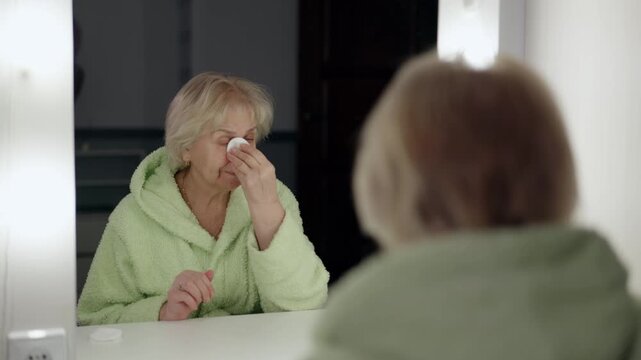 Mature woman wearing soft bathrobe sitting near vanity mirror, gently removing makeup using cotton pad during evening skincare routine, demonstrating personal beauty wellness