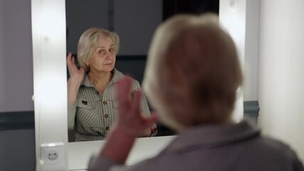Senior woman combing her hair while sitting in front of a mirror in a makeup room, checking her hairstyle and preparing for an important event