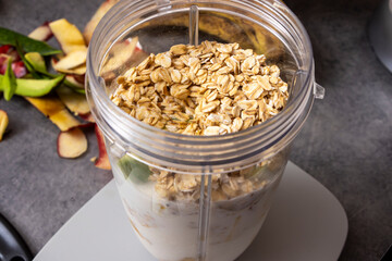 A blender jar filled with oats, yogurt, and fresh fruits sits on a kitchen countertop. Surrounding the jar are apple peels and slices, indicating meal prep and healthy eating.
