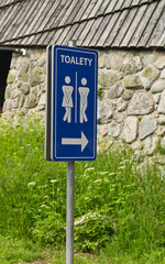 Public toilet direction sign with green grass and wall background providing clear guidance. Blue restroom sign with male and female icons standing near stone building in outdoor area during daylight.
