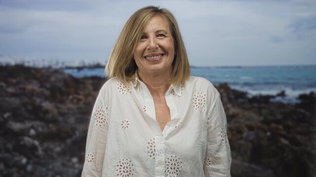 Woman smiles and tilts head in a white eyelet blouse at a rocky seaside beach under a cloudy sky; serenity.