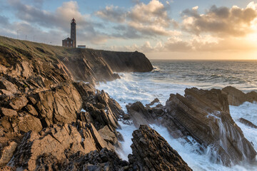 Coastal landscape with rough seas and dramatic geology. Sunset light with clouds. In the background, the Penedo da Saudade Lighthouse, in S&atilde;o Pedro de Moel, Portugal.