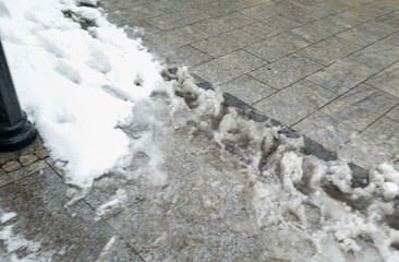 Melting snow creates mixed patches of ice and slush along a city sidewalk. Cold weather conditions illustrate the transition of seasons in urban landscapes.