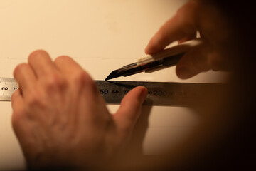 Over-the-shoulder view of hands cutting paper with a craft knife and ruler. Warm, focused light on a dark background highlights the meticulous process of creation and design.