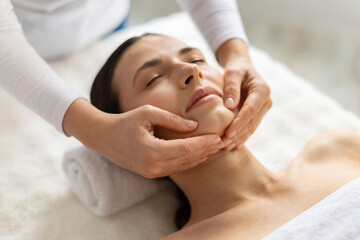 Young beautiful woman, female client receiving gentle chin massage at spa. Calm expression, warm light and skilled hands highlight beauty, skincare care, wellness and a peaceful lifestyle atmosphere