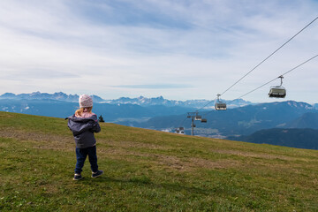A young girl, seen from behind, stands on a grassy alpine meadow on Gerlitzen mountain, gazing at the panoramic vista of a distant mountain range and a chairlift under a vast, cloudy sky in Austria.