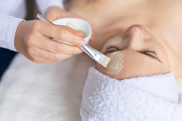 Young woman receiving precise clay mask application on her forehead. Clean spa environment, warm tones and relaxed posture highlight skincare care, facial treatment and calm wellness beauty