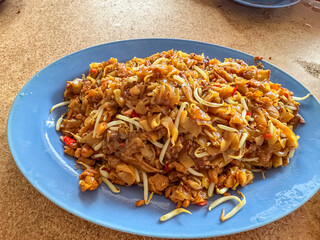 A close-up view of a freshly prepared plate of Char Kway Teow, a popular Malaysian stir-fried rice noodle dish with bean sprouts and chili, served on a blue plate at a local eatery in Melaka.