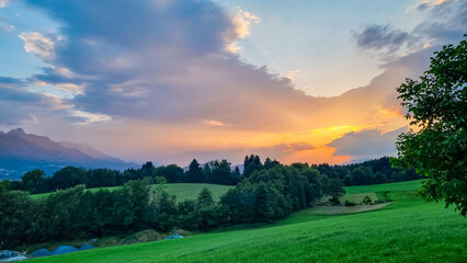 A dramatic golden sunset breaks through atmospheric clouds, casting beautiful, warm light over rolling green hills and forests in  rural Carinthian valley, with the Karawanken mountains in distance
