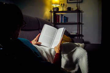 A woman wrapped in a blanket reads a book on the sofa in a dim living room, illuminated only by the warm glow of a floor lamp.