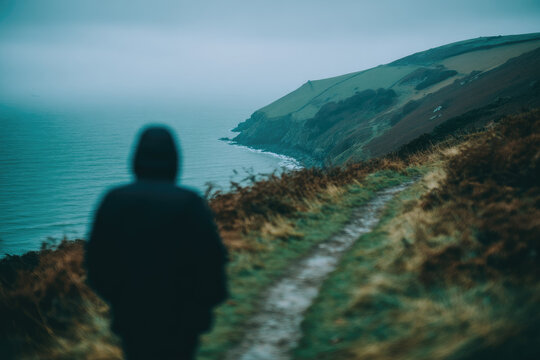 Defocused person walking on coastal path soft focus overcast sky ocean view rugged cliff grassy hillside tranquil moody atmosphere solitude nature