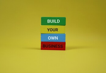 Yellow background highlights four wooden blocks spelling out 'BUILD YOUR OWN BUSINESS,' symbolizing entrepreneurship, self-made success, and creative startup energy in a bold, modern setting.