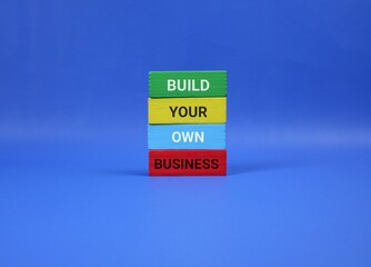 Blue background highlights four wooden blocks spelling out 'BUILD YOUR OWN BUSINESS,' symbolizing entrepreneurship, self-made success, and creative startup energy in a bold, modern setting.
