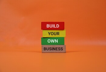 orange background highlights four wooden blocks spelling out 'BUILD YOUR OWN BUSINESS,' symbolizing entrepreneurship, self-made success, and creative startup energy in a bold, modern setting.
