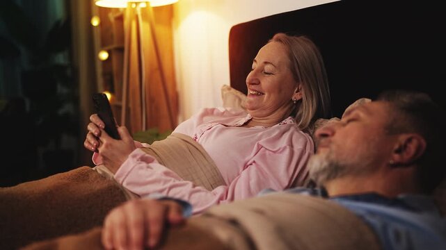 Woman in her 50s using dating app on phone while lying beside sleeping husband