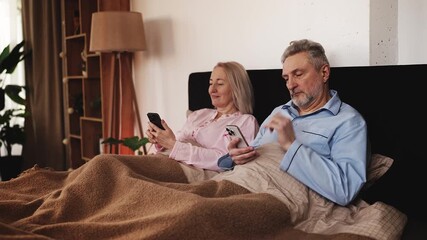 Married mature couple scrolling on phones while lying in bed, modern tech use - Powered by Adobe