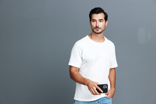 Confident young man standing against grey backdrop, holding smartphone, wearing casual white t-shirt and denim shorts. Modern lifestyle concept.