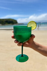 A hand holding a vibrant green cocktail with a lime garnish on a sandy beach. Bright tropical drink in a green glass, with the ocean and blue sky in the background.
